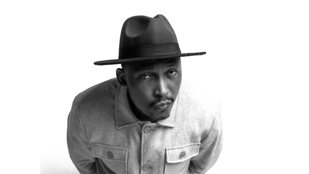 A high-angle, black and white studio portrait of Nigerian actor Frank Donga (Kunle Idowu). He is wearing a light-coloured button-up jacket and a dark fedora hat, looking up at the camera with a serious, deadpan expression against a plain white background.
