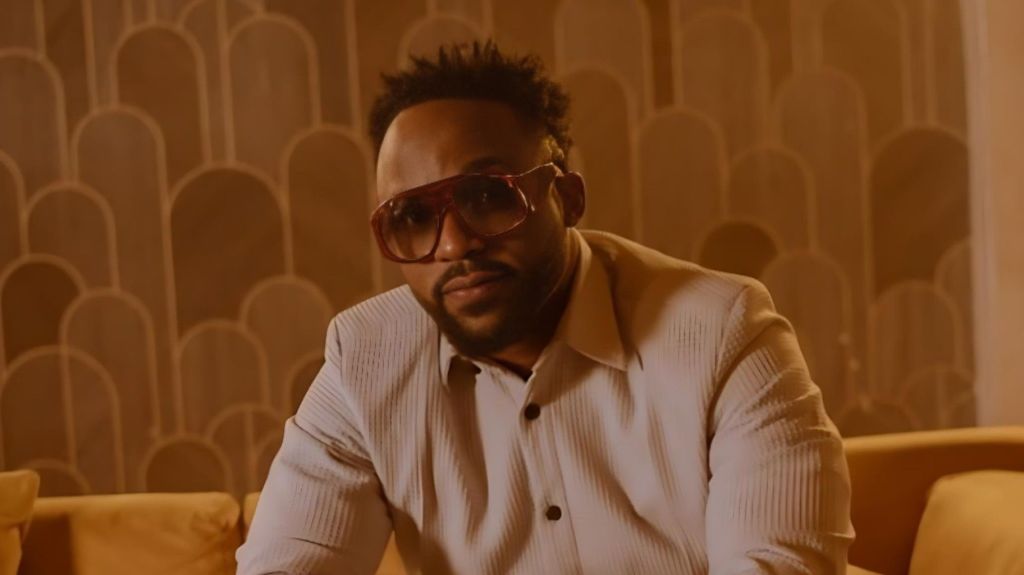 Nigerian singer Iyanya Mbuk sitting in a white shirt and tinted sunglasses against a patterned background.