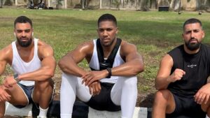 Photo of British heavyweight boxer Anthony Joshua (center: wearing white compression leggings, black shorts, white tank top, and a black weighted vest, sitting with arms on knees) flanked by two trainers on an outdoor grassy training ground—left trainer in white tank top and dark shorts with earbuds, right trainer in black sleeveless "KAMANI" shirt gesturing with hand.