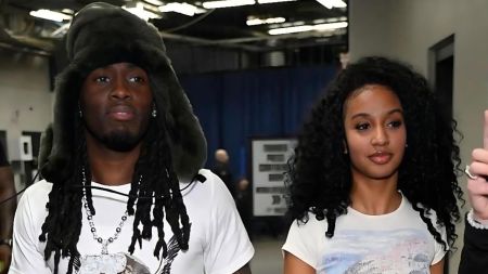 Gigi defends luxury demands from Kai Cenat and claims she drove Benz at 16 Kai Cenat and his ex-girlfriend Gigi standing side-by-side indoors. Kai, on the left, wears a white t-shirt, a silver chain, and a large, dark, furry trapper hat. Gigi, on the right, wears a white graphic t-shirt and has long, dark curly hair.