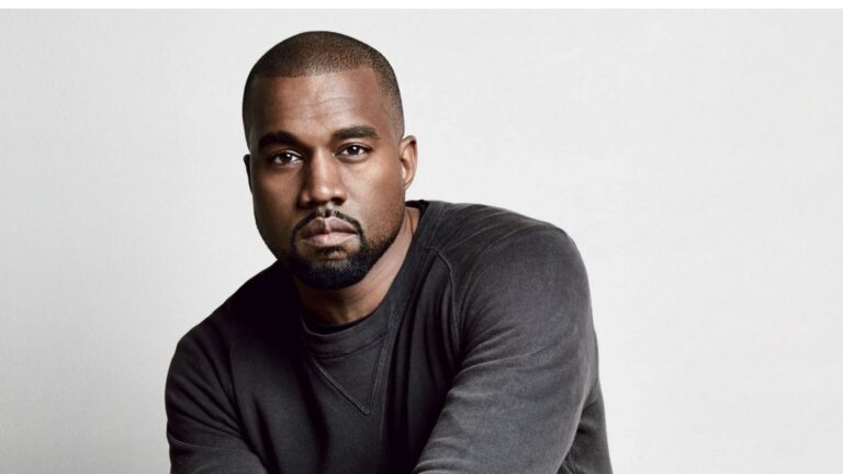 Studio portrait of Ye (Kanye West) wearing a black crewneck sweatshirt, leaning forward with a serious expression against a plain white background.