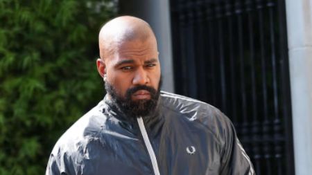 A close-up outdoor shot of Ye (Kanye West) standing in front of a black metal gate and greenery. He has a shaved head and a full dark beard. He is wearing a black Fred Perry track jacket with white piping details on the zipper and collar, looking off-camera with a serious expression.