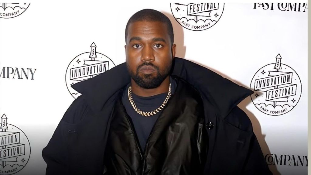 Kanye West (Ye) poses for a photo at the 2019 Fast Company Innovation Festival in New York City. He is wearing a black t-shirt, a gold chain necklace, and a large, oversized black coat with a high collar. The white backdrop behind him features a repeating pattern of "Fast Company" and "Innovation Festival" logos.