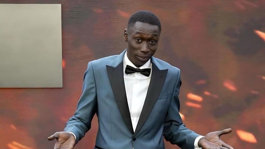 Khaby Lame stands on a red carpet at the 2025 Met Gala, wearing a custom grey BOSS tuxedo with black peak lapels, a white winged-collar shirt, and a black bow tie. He is looking directly at the camera with a neutral expression while performing his signature "shrug" hand gesture, palms facing upward. The background is a blurred warm brown, typical of the event's "Garden of Time" or "Superfine" thematic decor.