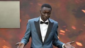 Khaby Lame stands on a red carpet at the 2025 Met Gala, wearing a custom grey BOSS tuxedo with black peak lapels, a white winged-collar shirt, and a black bow tie. He is looking directly at the camera with a neutral expression while performing his signature "shrug" hand gesture, palms facing upward. The background is a blurred warm brown, typical of the event's "Garden of Time" or "Superfine" thematic decor.