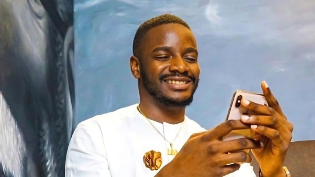 Leo DaSilva smiling while looking at his mobile phone, wearing a white native outfit with a gold brooch.