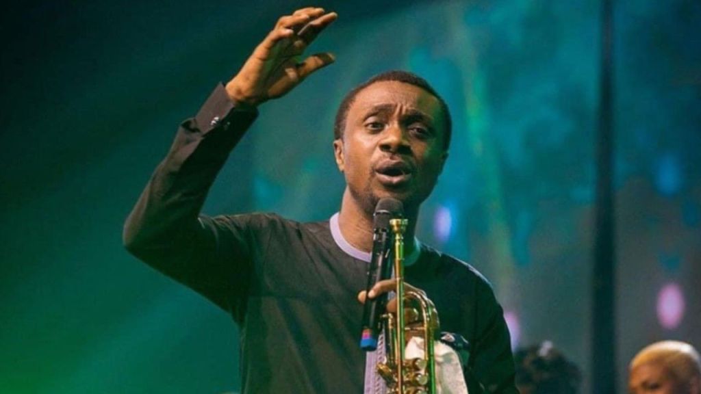 Nathaniel Bassey singing into a microphone with his right hand raised and holding a trumpet in his left hand during a stage performance with green lighting in the background.