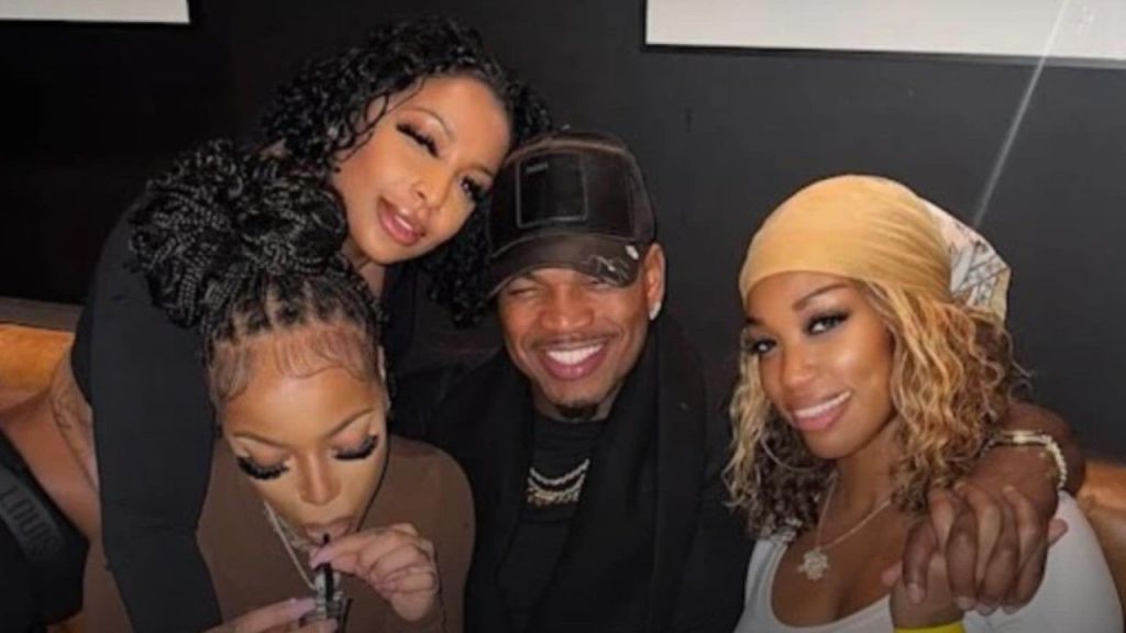 R&B singer Ne-Yo smiles in a close-up group photo with his three girlfriends; the woman on the left with dark curly hair leans on his shoulder, the woman below her drinks from a straw, and the woman on the right wears a beige headscarf and holds his hand.