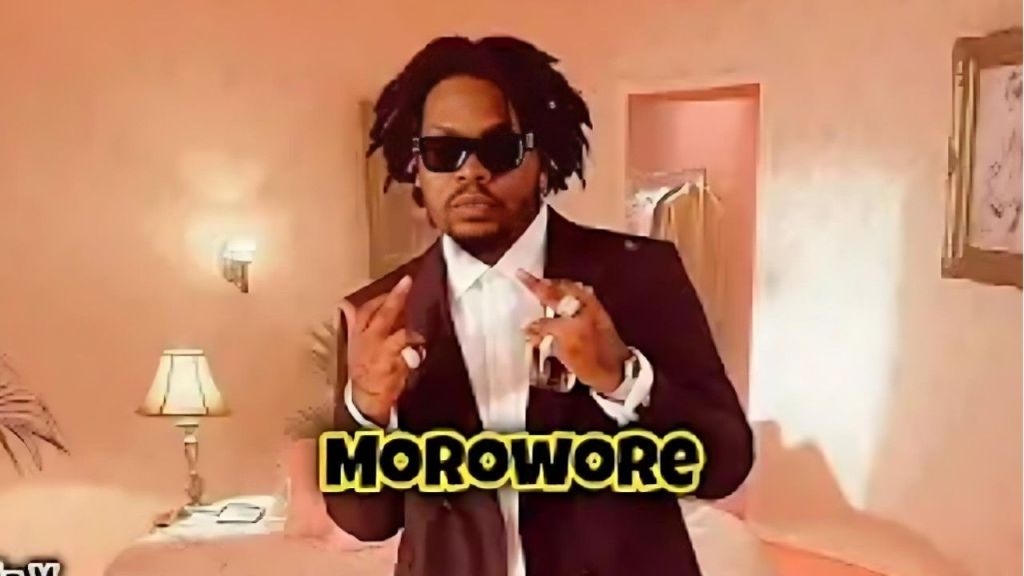 Nigerian rapper Olamide wearing a dark suit and sunglasses, gesturing with his hands in a room with warm pink lighting, featuring the text "MOROWORE" overlaid in yellow font at the bottom.
