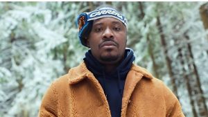 Nigerian singer Jaywon posing in a snowy forest environment. He is looking directly at the camera, wearing a brown textured shearling jacket over a dark blue hoodie, with Carrera brand ski goggles resting on his forehead.