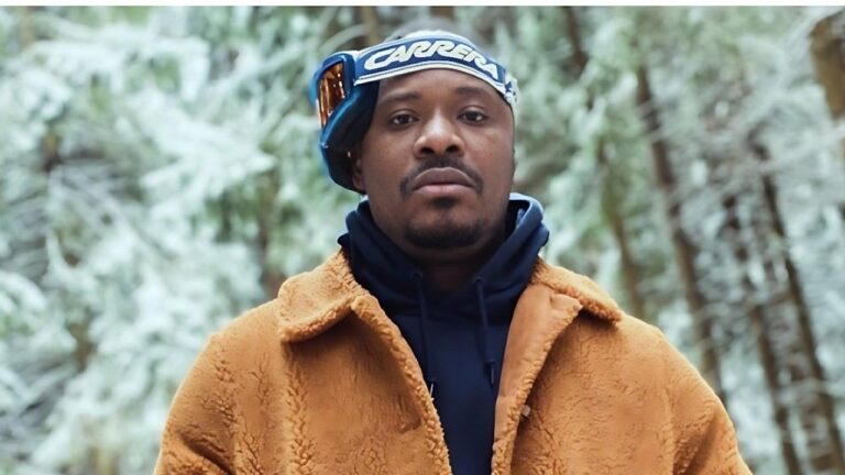Nigerian singer Jaywon posing in a snowy forest environment. He is looking directly at the camera, wearing a brown textured shearling jacket over a dark blue hoodie, with Carrera brand ski goggles resting on his forehead.