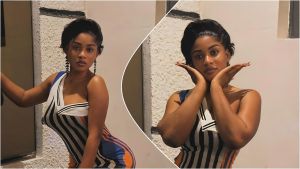 A split collage features two photos of BBNaija star Phyna posing in a fitted, sleeveless dress with black, white, blue, and orange vertical stripes. On the left, she stands against a white wall, looking seriously at the camera with one hand resting on a doorframe. On the right, in a circular insert, she frames her face with both hands, maintaining a calm, direct gaze. She wears dangling earrings and sports a short, sleek black hairstyle in both shots.