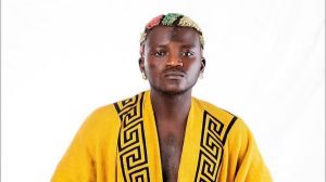 A studio portrait of Nigerian singer Portable (Habeeb Okikiola) looking directly at the camera with a serious expression. He is wearing a bright yellow robe featuring black Greek key patterns on the lapels and sports his signature multi-colored dyed hair against a plain white background.