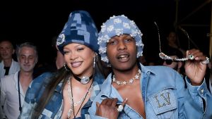 Close-up of Rihanna and A$AP Rocky at a fashion event. They are wearing matching blue pixelated-denim outfits and blue beanies; Rocky's beanie is embellished with pearls. He is holding a pair of sunglasses with pearl-encrusted frames in his left hand while they both smile at the camera.
