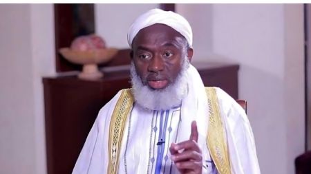 Sheikh Ahmad Gumi speaking during an interview, wearing a white turban and traditional white and gold robes while gesturing with his right hand.