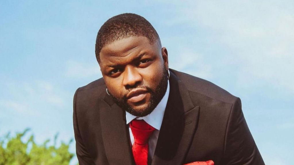 Nigerian music star Skales wearing a formal black suit, red tie, and silver hoop earring while posing outdoors against a blue sky.