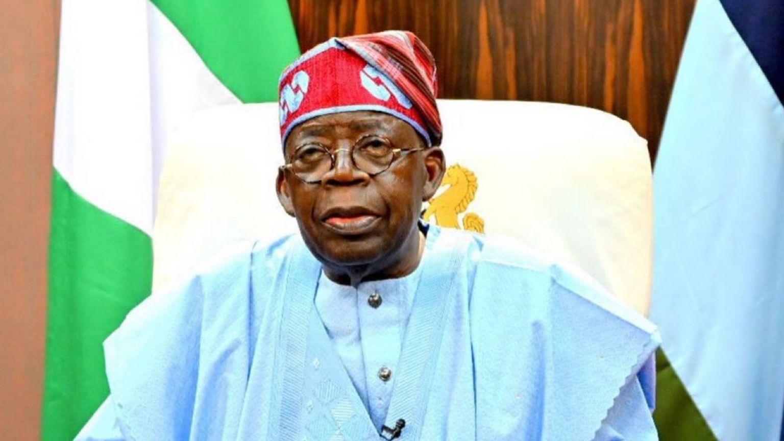 Official portrait of Nigerian President Bola Ahmed Tinubu seated in an executive office setting. He is wearing a light blue, embroidered agbada and his signature red cap with blue geometric embroidery. He is also wearing glasses. The green-white-green Nigerian national flag is visible in the background behind his right shoulder, alongside the flag of the Commander-in-Chief.