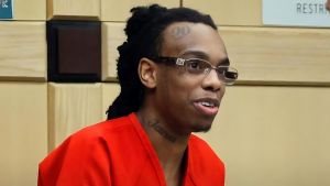 YNW Melly (Jamell Demons) appearing in court wearing an orange prison jumpsuit and glasses. He is smiling and looking to the side, with visible tattoos on his forehead ("90") and neck ("Jana").