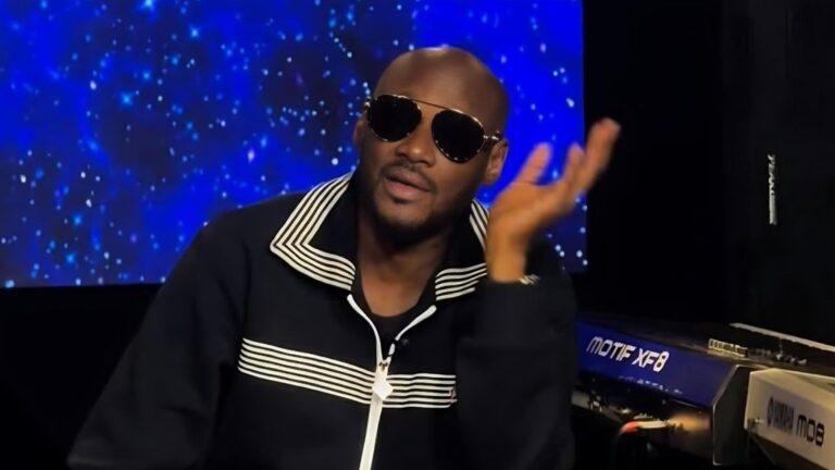 A close-up of Nigerian singer 2baba wearing dark aviator sunglasses and a black zip-up jacket with white horizontal stripes across the chest and collar. He is gesturing with his left hand while seated in front of a blue starlight-themed background and a Motif XF8 keyboard.