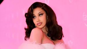 A portrait of American rapper Cardi B with voluminous, dark auburn wavy hair. She is wearing an off-the-shoulder light pink gown with delicate feather detailing along the neckline. Her makeup features a bold winged eyeliner and a soft pink lip, set against a solid pink background.
