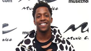 A head-and-shoulders portrait of American rapper ILoveMemphis (also known as iHeartMemphis) smiling brightly. He has short, dark curly hair and is wearing a black torn shirt under a white jacket with large black polka dots. The background features multiple repeating logos for "Music Choice".