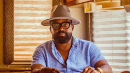 Nollywood filmmaker Kunle Afolayan seated indoors, wearing a brown fedora hat, black-rimmed glasses, and a light blue short-sleeved shirt.
