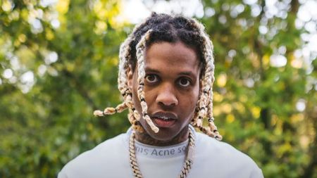 Rapper Lil Durk wearing a white Acne Studios shirt and gold chains while posing in front of a background of green trees.