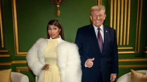 Nicki Minaj, wearing a yellow dress and large white fur coat, stands next to Donald Trump, who gives a thumbs up in a room with green and gold walls.
