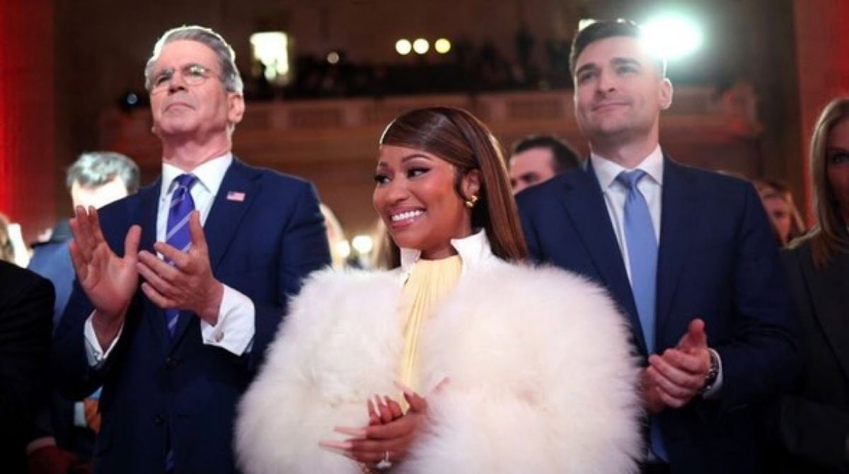 Nicki Minaj smiling and clapping while wearing a white fur coat, standing between two men in blue suits at a formal event.