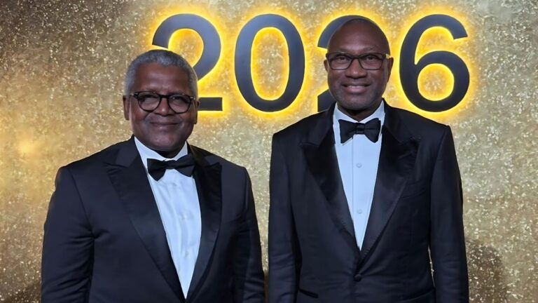 A professional photograph shows Nigerian billionaires Aliko Dangote (left) and Femi Otedola (right) standing side-by-side. Both men are smiling and dressed in formal black tuxedos with bowties. They are posed in front of a shimmering gold textured backdrop that features a large, illuminated "2026" sign directly between them.