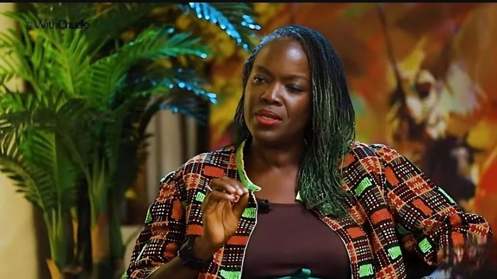 A medium shot of Eyiyemi "Yemi" Pratt, a veteran Nigerian broadcaster and fashion designer, during an interview on the show #WithChude. She is a Black woman with dark hair styled in long, thin micro-braids with green-tinted ends. She is wearing a dark brown top under an open jacket featuring a vibrant, multicolored African print with geometric patterns in orange, red, green, and black. Her expression is thoughtful and engaged as she gestures with her right hand while speaking against a warm, blurred background featuring indoor plants and abstract art