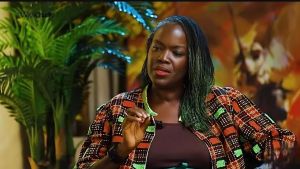 A medium shot of Eyiyemi "Yemi" Pratt, a veteran Nigerian broadcaster and fashion designer, during an interview on the show #WithChude. She is a Black woman with dark hair styled in long, thin micro-braids with green-tinted ends. She is wearing a dark brown top under an open jacket featuring a vibrant, multicolored African print with geometric patterns in orange, red, green, and black. Her expression is thoughtful and engaged as she gestures with her right hand while speaking against a warm, blurred background featuring indoor plants and abstract art