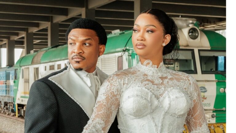 A cinematic medium close-up of Nollywood actor Shawn Faqua and his bride, Sharon Ifunanya, posing in their white wedding attire. Shawn wears a modern black tuxedo with unique white pearl-beaded lapels and a white cravat, looking off to the left. Sharon stands slightly behind him, dressed in a high-neck, long-sleeved white lace gown with a pearl-embellished bodice, looking toward the right. In the background, a green and white Nigerian Railway Corporation (NRC) locomotive is visible at the Mobolaji Johnson Train Station, marking Nigeria's first-ever wedding ceremony held on a moving train.