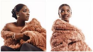 Two side-by-side portraits of Nigerian artist Simi against a bright white background. In both, she is wrapped in a voluminous, textured copper-brown faux fur coat. On the left, she is seen in profile with a sleek low bun and a bold, square gold earring, looking off-camera with a serene expression. On the right, she faces slightly forward with a playful, open-mouthed expression, her dark hair middle-parted and slicked back. The lighting is soft and high-key, emphasizing her radiant skin and the rich texture of the fur.