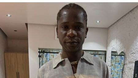 Close-up photo of Nigerian rapper Speed Darlington standing indoors. He is wearing a grey collared shirt with a gold chain necklace and has a serious expression. A wardrobe and a unique mirror are visible in the background.