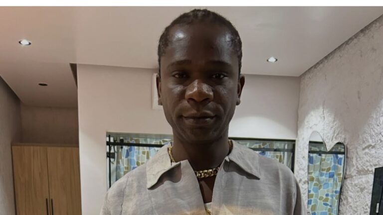 Close-up photo of Nigerian rapper Speed Darlington standing indoors. He is wearing a grey collared shirt with a gold chain necklace and has a serious expression. A wardrobe and a unique mirror are visible in the background.